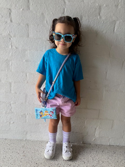 A young girl wearing a B.Balanced Co. blue crew neck t-shirt with pink shorts, sunglasses, and a small crossbody bag.