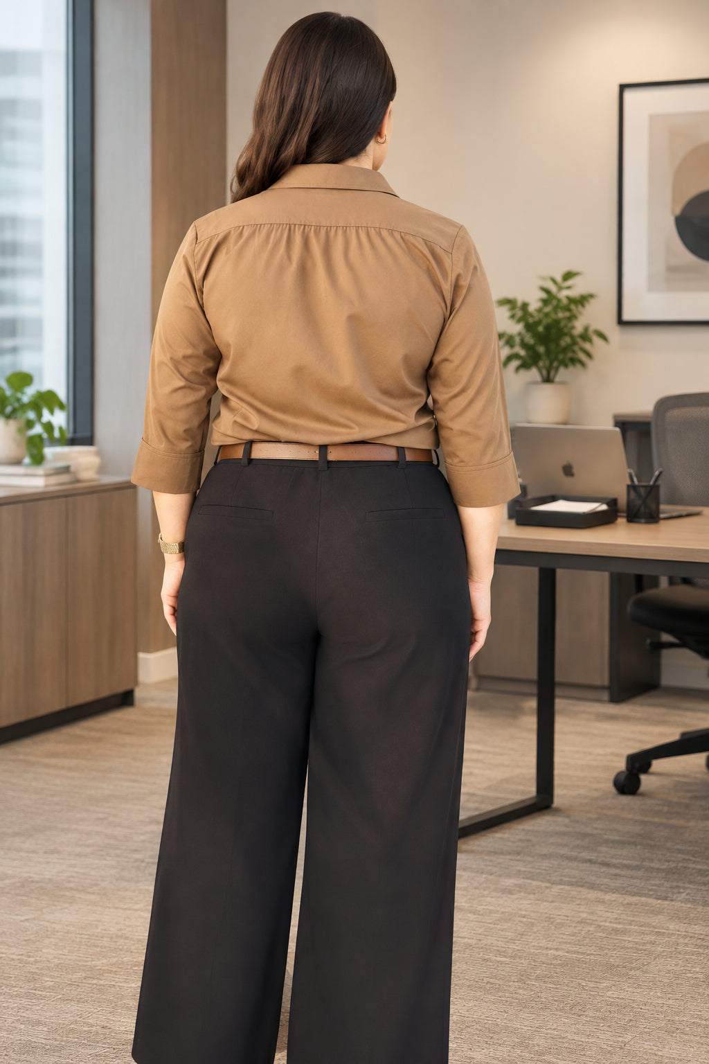 A woman viewed from behind wearing a tan long-sleeve button-up shirt and dark charcoal trousers, standing in front of a corrugated metal fence.