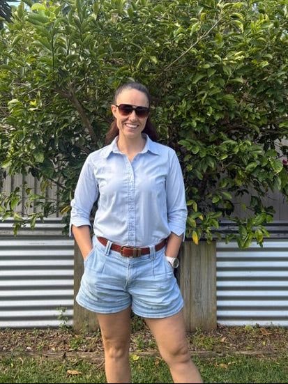 A women standing in front of a tree wearing a light blue shirt with shorts and a brown belt 
