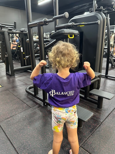 Child wearing a purple B.Balanced Co. Balanced Kids crew neck t-shirt with back logo in a gym setting