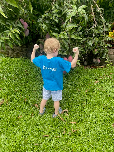 Toddler wearing a bright blue B.Balanced Co. crew neck t-shirt with logo, standing in a garden by a mango tree.