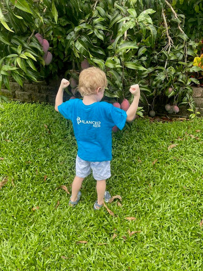 Toddler wearing a bright blue B.Balanced Co. crew neck t-shirt with logo, standing in a garden by a mango tree.