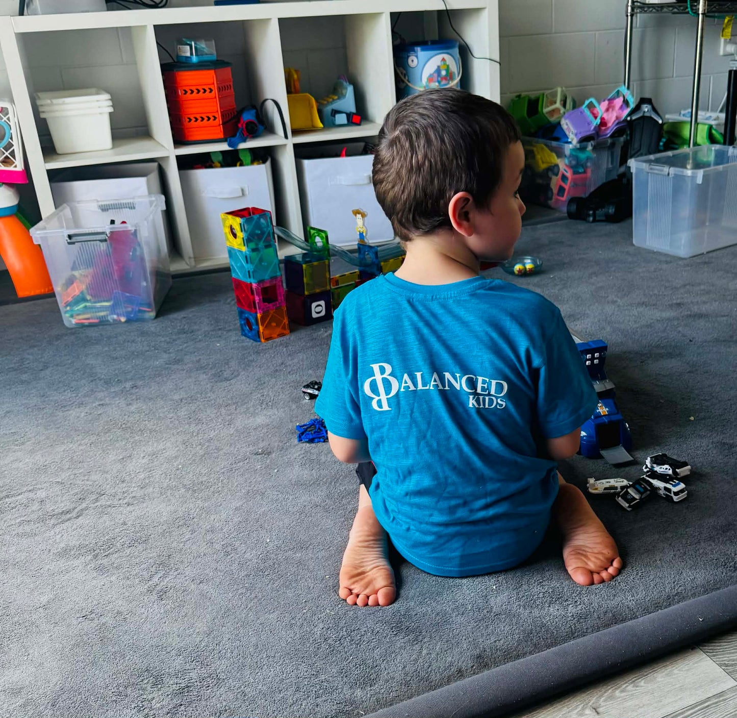 A young boy wearing a bright blue B.Balanced Co. crew neck t-shirt with Balanced Kids logo, playing on a grey rug.