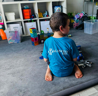 A young boy wearing a bright blue B.Balanced Co. crew neck t-shirt with Balanced Kids logo, playing on a grey rug.