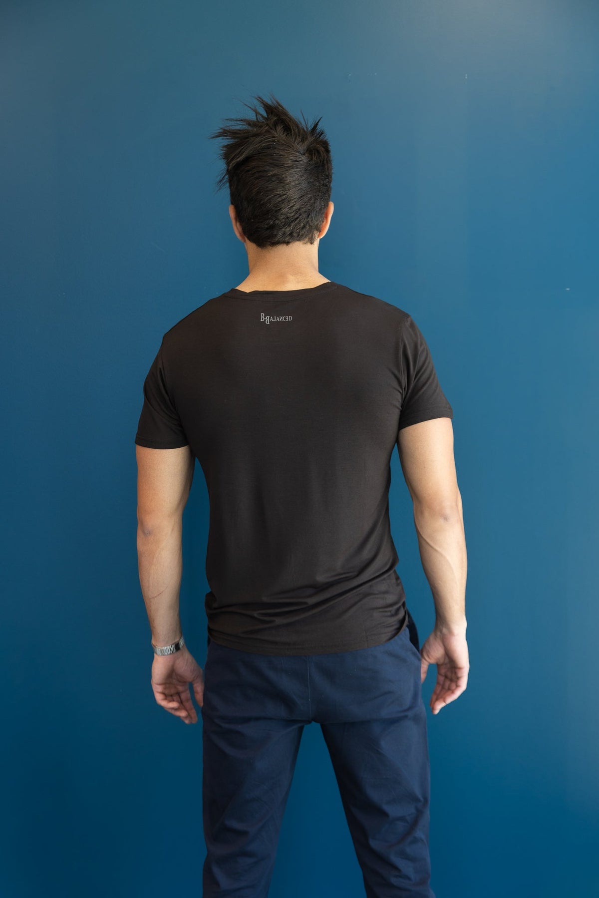 Back view of a man wearing a B.Balanced Co black bamboo blend Everyday Tee against a blue wall.