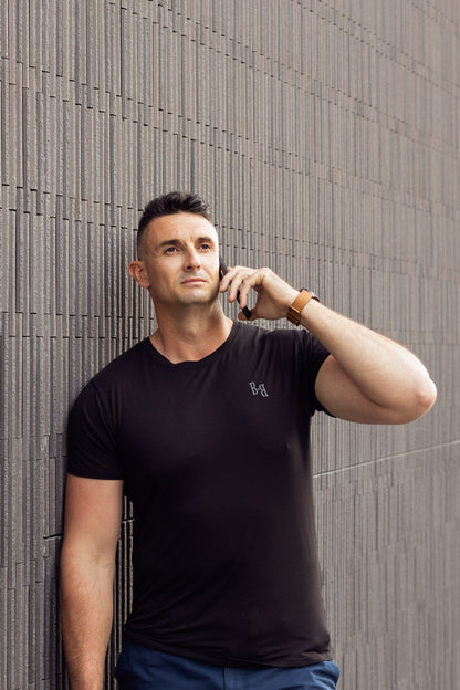 A man wearing a B.Balanced Co black bamboo blend Everyday Tee, leaning against a textured grey wall while on a call.