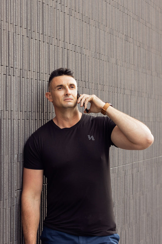 A man wearing a B.Balanced Co black bamboo blend Everyday Tee, leaning against a textured grey wall while on a call.
