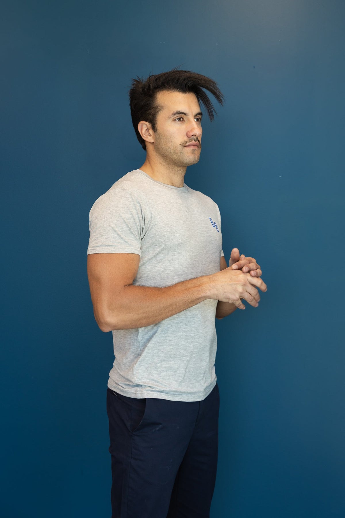 Man wearing a B.Balanced Co heathered grey crew neck t-shirt with blue logo, standing against a dark blue background.