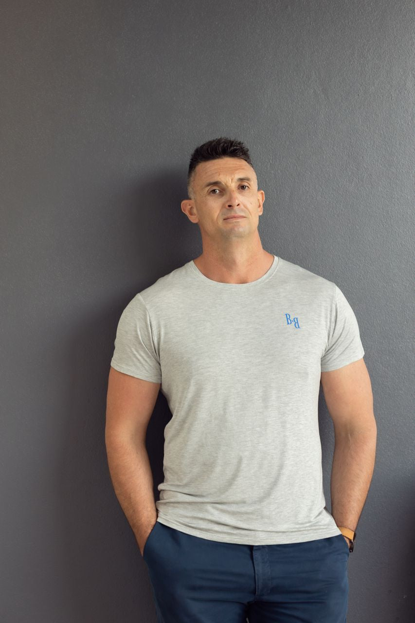 Man wearing a B.Balanced Co heathered grey crew neck t-shirt with blue logo detail against a dark grey wall