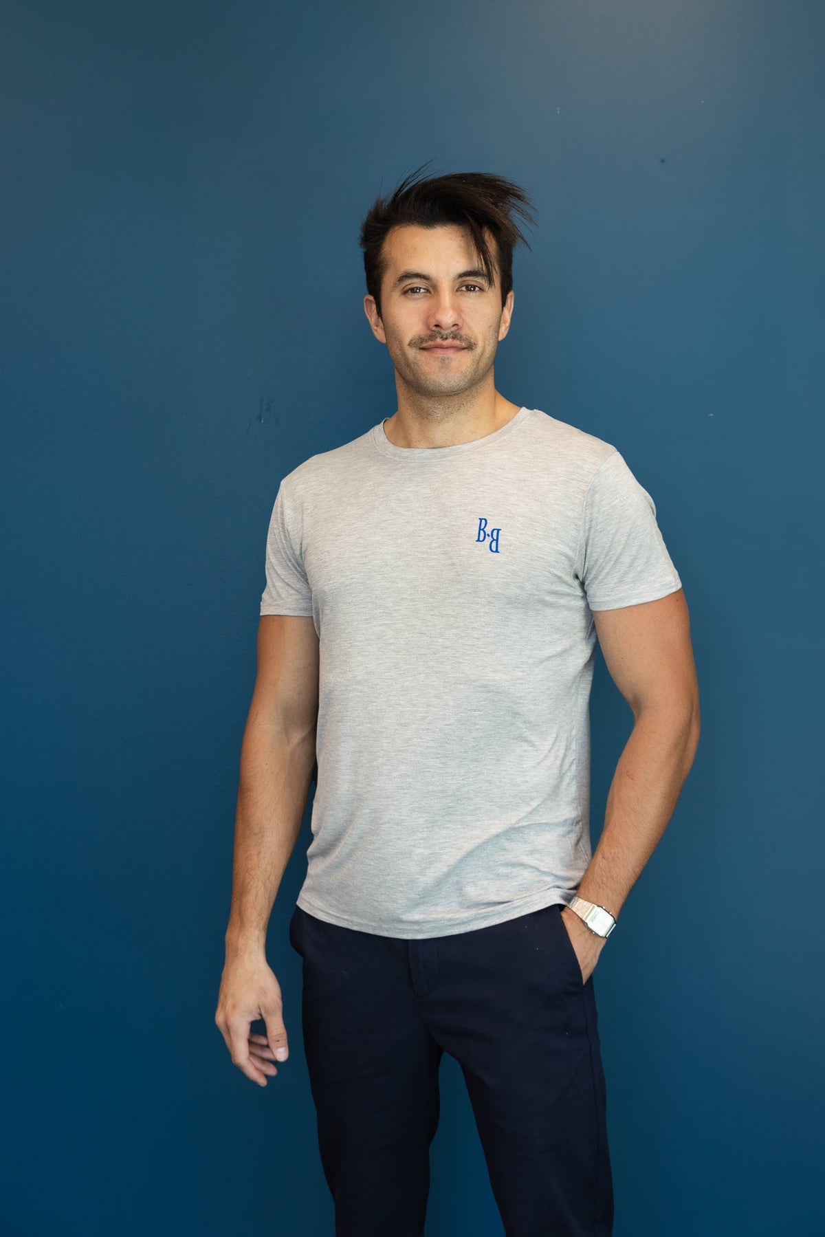 Man wearing a B.Balanced Co heathered grey crew neck t-shirt with blue chest branding against a blue wall