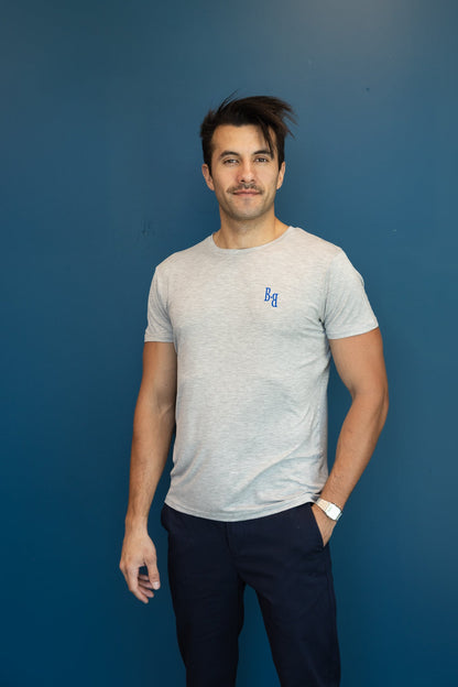 Man wearing a B.Balanced Co heathered grey crew neck t-shirt with blue chest branding against a blue wall